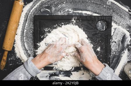 deux mains de femmes pétrient la pâte de farine de blé blanc sur une table noire, vue de dessus Banque D'Images