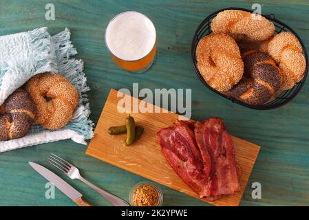 En-cas à base de viande, tranches de bacon et cornichons grillés servis sur une planche à découper en bois, moutarde grossière, pain et un verre de bière non filtrée. Banque D'Images