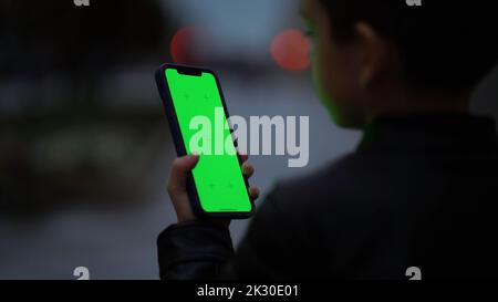 Garçon tapotant sur l'écran vert de son smartphone. Un enfant avec un téléphone dans ses mains regarde la chromey la nuit à l'automne. Banque D'Images