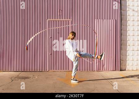 Woman standing on one leg playing with rainbow colored gymnastics ribbon in front of corrugated wall Stock Photo