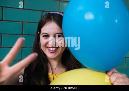 Une femme heureuse faisant signe de paix tenant des ballons bleus et jaunes Banque D'Images