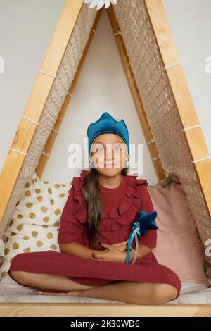 Jolie fille portant la couronne tenant la baguette magique assise dans la tente à la maison Banque D'Images