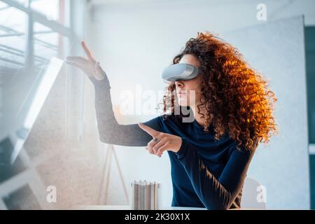 Jeune ingénieur aux cheveux bouclés portant un simulateur de réalité virtuelle assis au bureau Banque D'Images