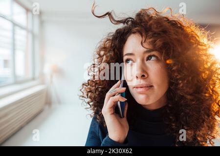 Belle femme aux cheveux bouclés qui parle sur un téléphone intelligent en contemplant Banque D'Images