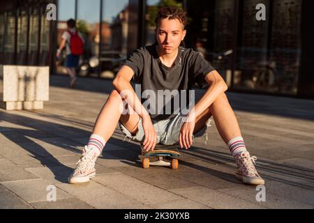 Confident non-binary person sitting on skateboard at footpath Stock Photo