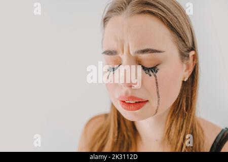 Sad young woman with smudged mascara crying in front of wall Stock Photo