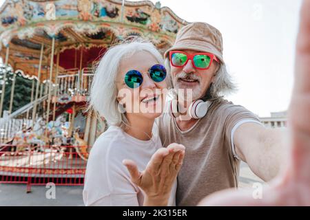 Homme âgé avec une femme adulte prenant le selfie devant le carrousel au parc d'attractions Banque D'Images