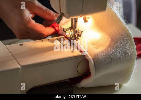 Les mains d'un travailleur expérimenté dans l'industrie artisanale, la couture de tissus rouges et blancs sur une machine à coudre, la production de chapeau rouge de Santa. Cousu maison Banque D'Images