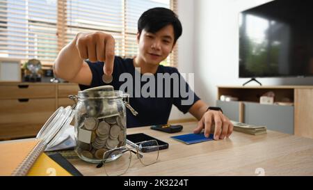 Homme asiatique souriant mettant la pièce dans le pot en verre, économiser de l'argent pour l'avenir, la retraite et le concept de planification financière Banque D'Images