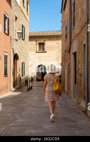 Femme vue de derrière, avec un chapeau, un sac et une robe colorée, marchant le long d'une rue typique dans la ville Majorcan d'Alcudia, le matin ensoleillé d'été Banque D'Images