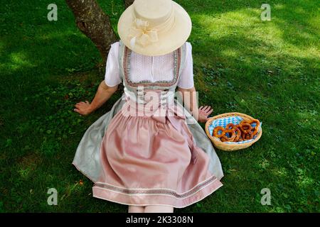 Une femme dans une belle robe traditionnelle bavaroise de dirndl assis sur la prairie verte avec un panier de bretzels ou Brezeln (Munich, Bavière) Banque D'Images