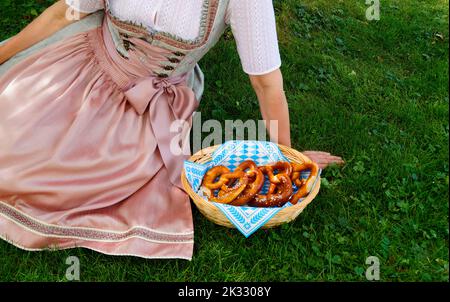 Une femme dans une belle robe traditionnelle bavaroise de dirndl assis sur la prairie verte avec un panier de bretzels ou Brezeln (Munich, Bavière) Banque D'Images