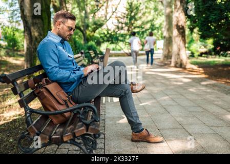 Homme adulte concentré, dactylographiant sur son ordinateur portable, travaillant au parc, prenant son travail au sérieux. Banque D'Images
