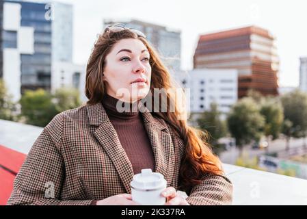 Femme branchée et attentionnés avec une tasse de café à emporter à emporter sans aucune perte tout en se reposant sur un arrière-plan flou de la rue de la ville en journée Banque D'Images