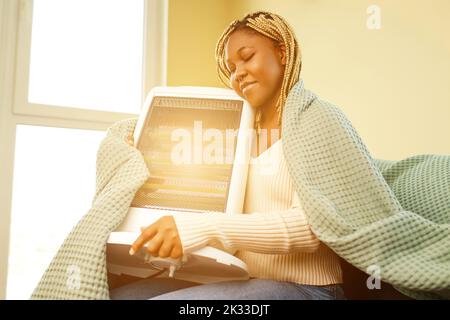 Femme afro-américaine dans une couverture bleue se réchauffant près d'un chauffage électrique à la maison Banque D'Images