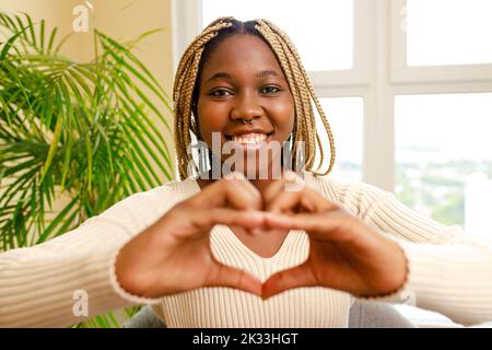 femme hispanique montrant l'amour avec les mains en forme de coeur exprimant le symbole de santé et de mariage près de la fenêtre et la plante verte à la maison Banque D'Images