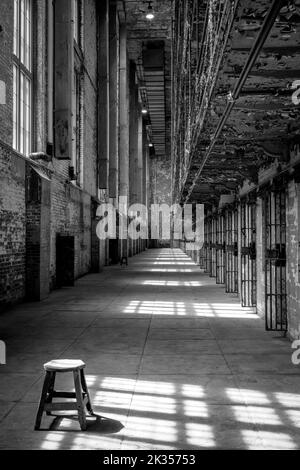 Tabouret assis dans le couloir d'une prison abandonnée, avec des ombres sur le sol des bars dans les fenêtres. Image noir et blanc (noir et blanc). Banque D'Images