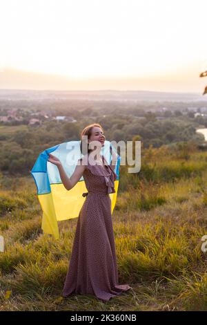 Femme heureuse en robe longue portant un drapeau jaune et bleu de l'Ukraine qui flotte dans le vent sur un fond de coucher de soleil. Les Ukrainiens contre la guerre. Inde Banque D'Images