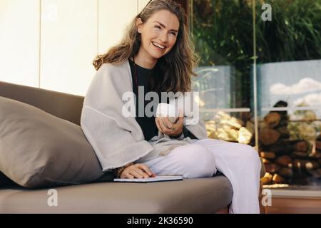 Une femme âgée gaie qui fait un journal avec une tasse de thé le matin. Femme mûre souriant en s'asseyant sur un canapé. Femme sénior se détend Banque D'Images