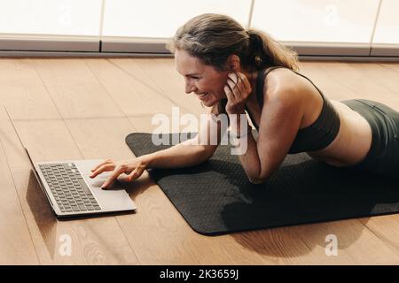 Femme âgée souriant tout en participant à un cours de fitness en ligne sur un ordinateur portable. Bonne femme suivant un tutoriel de yoga en ligne à la maison. Femme âgée gaie l Banque D'Images