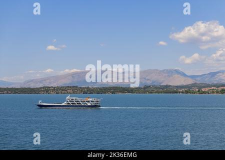 Le petit ferry local bleu et blanc traverse la baie de Garitsa sur la toile de fond des hautes collines du centre de l'île de Corfou avec une côte boisée, Banque D'Images