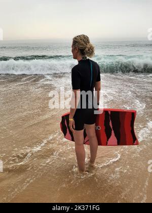 Femme blonde en combinaison debout sur la plage avec planche de surf Banque D'Images