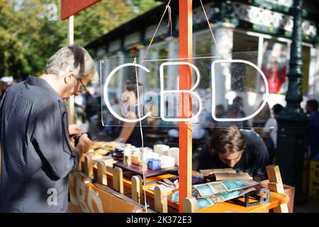 Man regarde les produits CBD de la marque 'Kadra' sur un stand vendant une gamme de produits CBD (cannididiol) en dehors du café Rosa Bonheur, lors d'une journée ensoleillée au Parc des Buttes Chaumont, Paris, France Banque D'Images