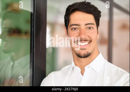 Joyeux et serein homme d'affaires latin dans une chemise blanche formelle, tête de jeune homme beau, ouvrier de collier blanc de sexe masculin regarde la caméra avec un sourire enivant Banque D'Images