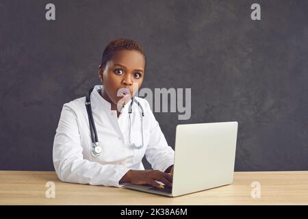 Femme médecin à la peau foncée avec un visage choqué assis à la table sur un fond gris. Banque D'Images