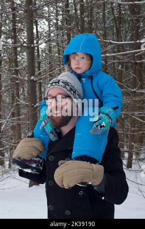 Un homme barbu dans un chapeau d'hiver donne à son todder adapté à la neige une promenade à l'épaule dans les bois enneigés canadiens. Banque D'Images