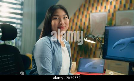 Femme asiatique 3D designer assis à la table près de l'ordinateur moderne et de la tablette numérique, travaillant sur 3D projet à distance de la maison et regardant l'appareil photo. Banque D'Images