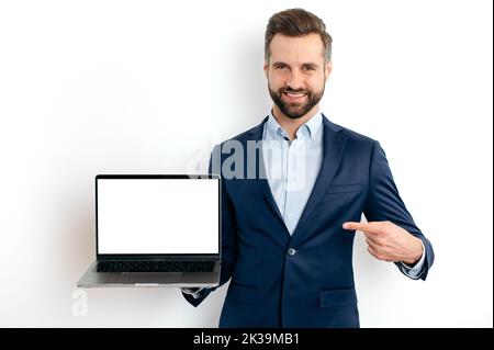 Homme d'affaires caucasien positif, en costume, programmeur, spécialiste INFORMATIQUE, tenant un ordinateur portable ouvert avec écran de maquette vide en main, pointe le doigt dessus, debout sur un arrière-plan blanc isolé, sourit Banque D'Images