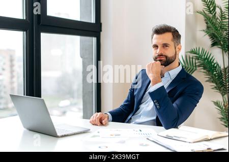 Photo d'un beau et positif élégant homme caucasien, entrepreneur, pdg de l'entreprise, assis au bureau, vêtu d'un costume habillé, regardant la caméra. Un homme d'affaires barbu et attrayant Banque D'Images