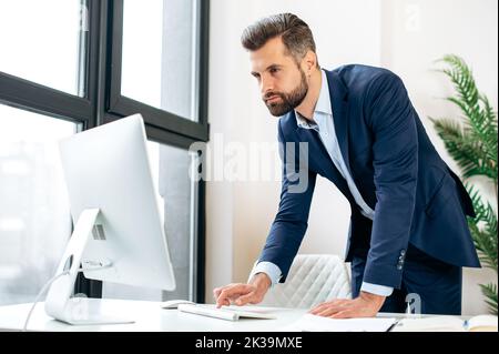 Occupé concentré confiance caucasien barbu homme, pdg de la société, directeur, stand près du bureau de travail dans le bureau, travail dans l'ordinateur, conduit la correspondance de travail en ligne, affiche des informations Banque D'Images