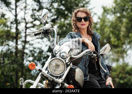 Portrait d'une femme mûre de rock glam debout à côté de sa moto et regardant l'appareil photo Banque D'Images