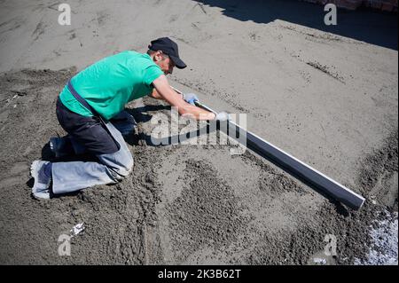 Un ouvrier pose le rail de table sur le sol recouvert de mélange sable-ciment sur le chantier de construction. Homme nivelant la surface avec bord droit tout en criant le sol à l'extérieur sur le toit du nouveau bâtiment. Banque D'Images