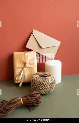 Boîte-cadeau et rouleau d'emballage, enveloppe, bougie sur table kaki. Fond de mur rouge. Anniversaire, anniversaire, noël, concept de salutation Banque D'Images