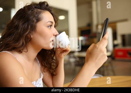 Profil d'une femme qui boit du café et vérifie son téléphone portable dans un bar Banque D'Images