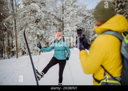 Homme senior prenant la photo de sa femme posant avec des skis dans la nature hivernale. Banque D'Images