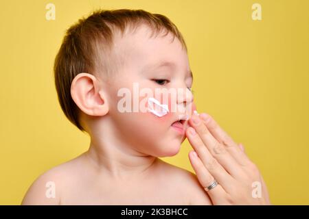 La mère sent la crème allergique sur le visage du bébé tout-petit, fond jaune studio. Portrait en gros plan d'un bébé mignon avec de la crème sur ses joues. Enfant âgé d'un an Banque D'Images