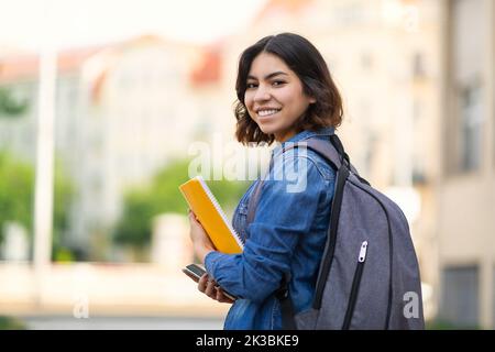 Jeune étudiante arabe souriante avec des livres de travail et un sac à dos debout à l'extérieur Banque D'Images