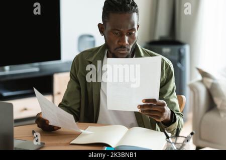 Homme afro-américain concentré de vérifier les documents, de calculer les factures et les taxes à l'aide d'un ordinateur portable dans l'intérieur de la chambre Banque D'Images