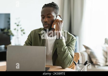 Homme indépendant afro-américain sérieux parlant sur téléphone portable et regardant ordinateur portable, travaillant en ligne à partir de la maison Banque D'Images