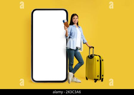 Gaie jeune femme chinoise dans une ambiance décontractée avec passeport de valise et billets d'avion près d'un immense smartphone avec écran vierge Banque D'Images