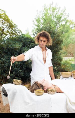 Le maître de bols de chant tibétain faisant le massage de guérison sonore avec le Népal Buddha chantant bol en plein air, jeune femme se détendre avec la médecine alternative. Un homme dedans Banque D'Images