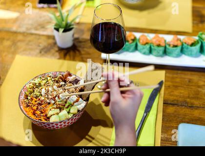 Avoir un plat de pique dans un restaurant. Un plat servi dans un bol avec sauce et garniture au sésame. Un verre de vin rouge et des petits pains à sushis sur la table du bac Banque D'Images