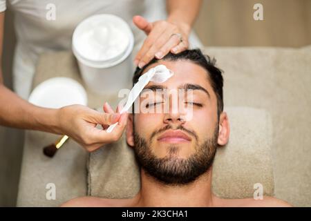Vue de dessus de la femme anonyme appliquant le masque purifiant sur le visage de jeune homme barbu client pendant le travail dans le salon de beauté Banque D'Images