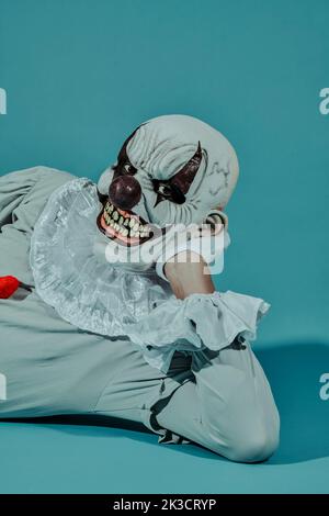 gros plan d'un clown mal à tête blanche et crépie, portant un costume gris avec une robe blanche, allongé sur le côté, penché sur un bras, sur un fond bleu Banque D'Images