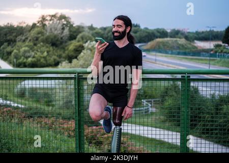 Jeune homme avec une jambe prothétique utilisant son smartphone pour communiquer au coucher du soleil Banque D'Images