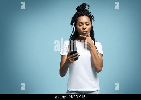 Une femme ethnique aux cheveux mauriques positifs utilise un téléphone portable vérifie les messages et lit les nouvelles tient le cellulaire moderne dans les mains regarde avec curieux heureux expression Banque D'Images